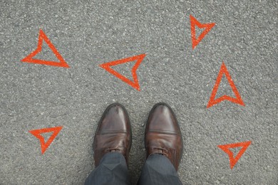 Image of Man standing near orange arrows pointing in different directions on asphalt road, closeup. Concepts of choice, decision, alternative way