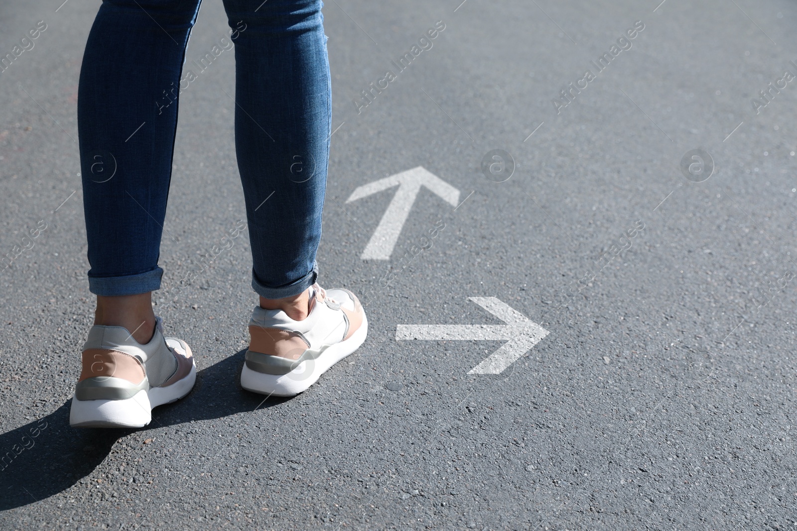 Image of Woman walking on asphalt road with white arrows pointing in different directions, closeup. Concepts of choice, decision, alternative way