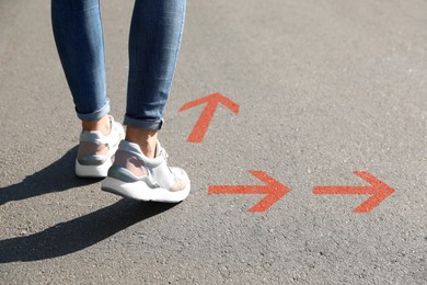 Image of Woman walking on asphalt road with orange arrows pointing in different directions, closeup. Concepts of choice, decision, alternative way