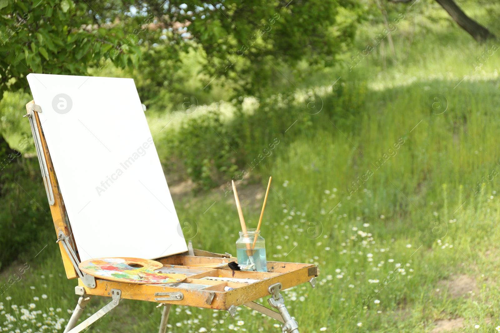 Photo of Easel with blank canvas and painting equipment in picturesque countryside