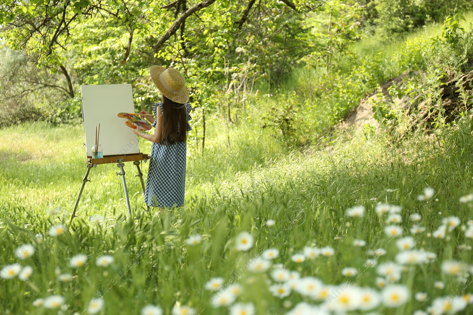 Photo of Little girl painting on easel in picturesque countryside, back view