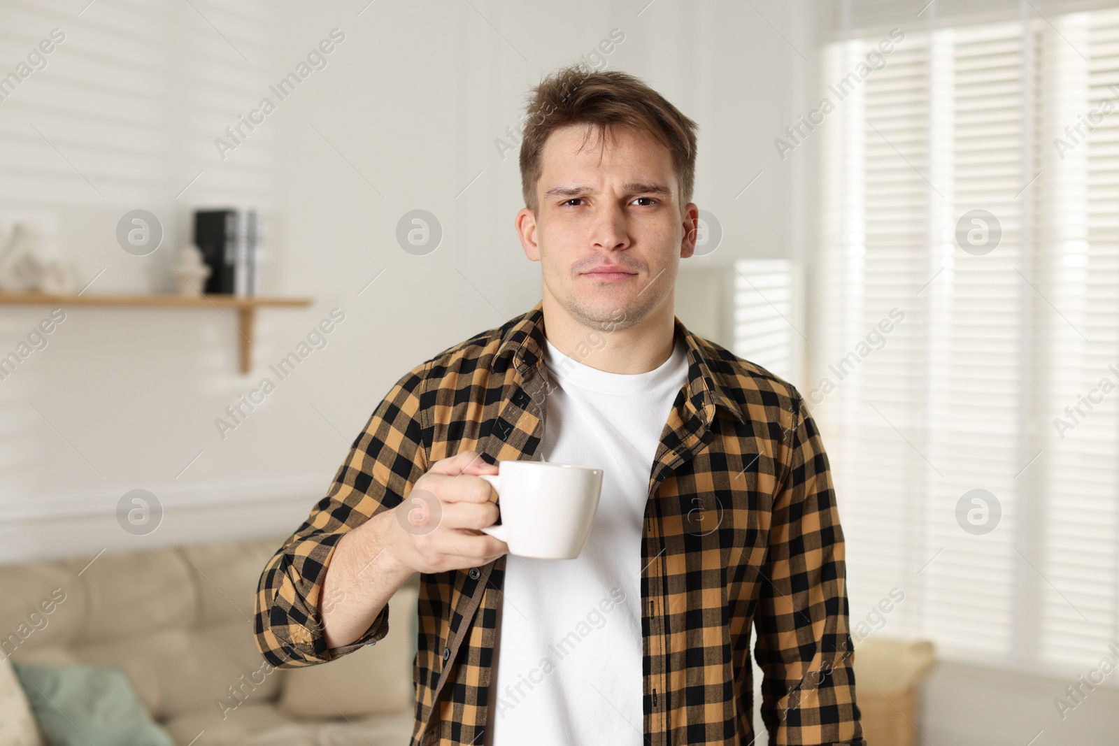 Photo of Unhappy young man with cup of coffee suffering from hangover at home