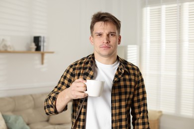 Photo of Unhappy young man with cup of coffee suffering from hangover at home