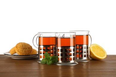 Glasses of aromatic tea in holders, cookies, lemon and mint on wooden table against white background