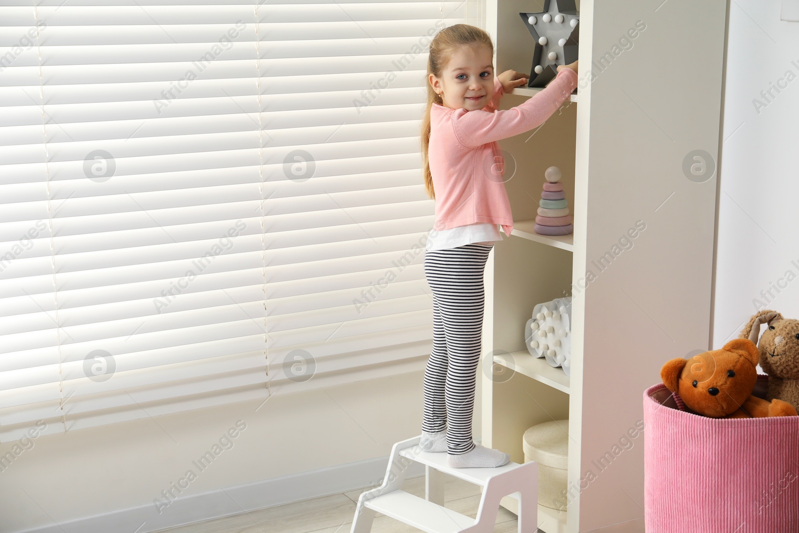 Photo of Little girl standing on step stool and reaching for toys on shelf indoors. Space for text