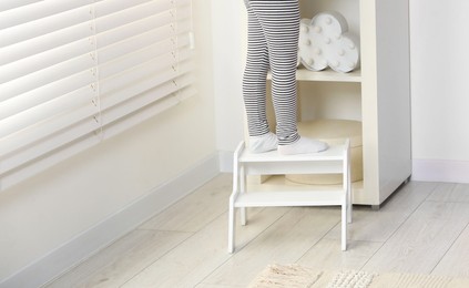 Photo of Little girl standing on step stool indoors, closeup. Space for text