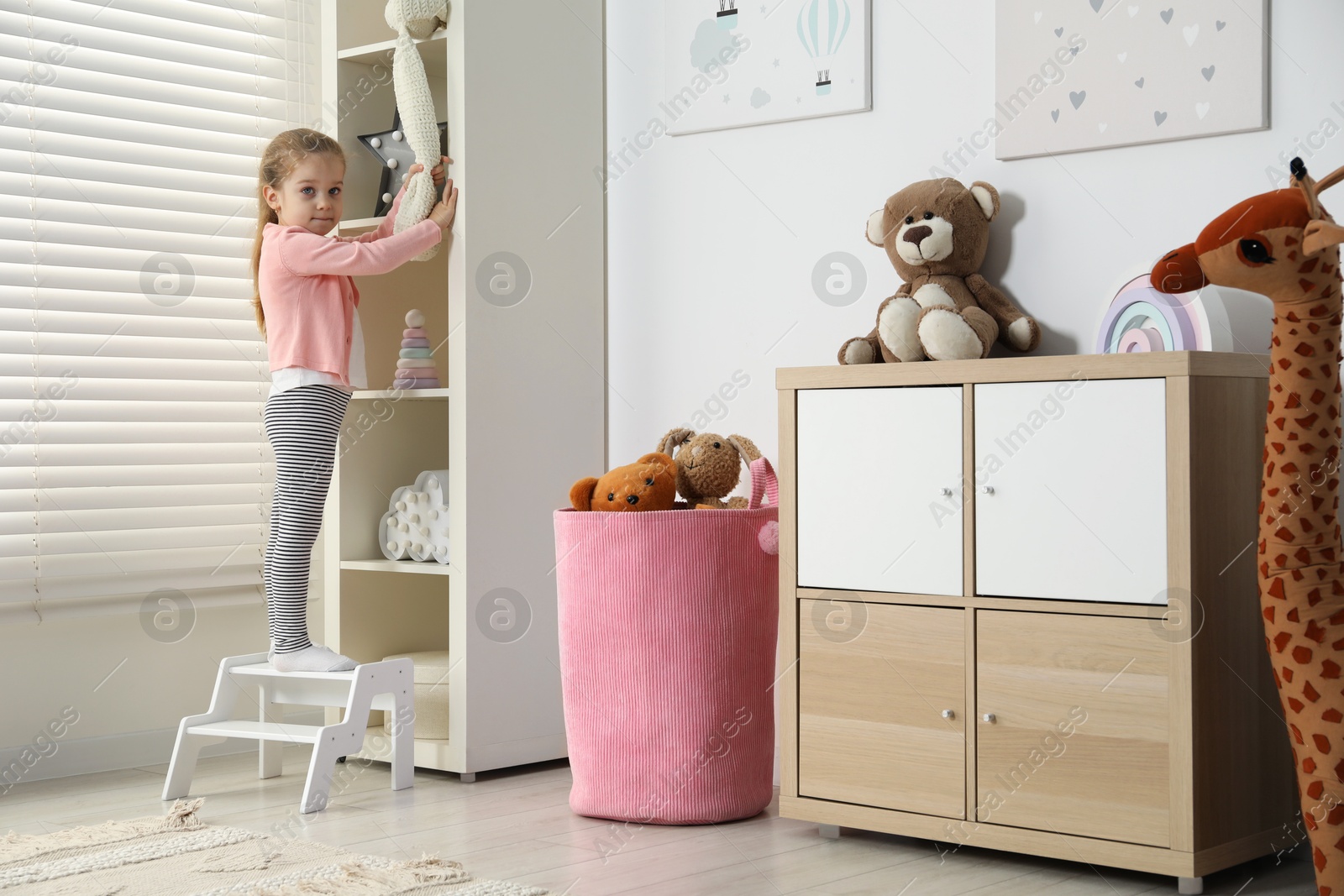 Photo of Little girl standing on step stool and reaching for toys on shelf indoors