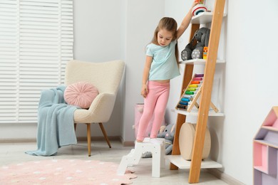 Photo of Little girl standing on step stool and reaching for toys on shelf indoors