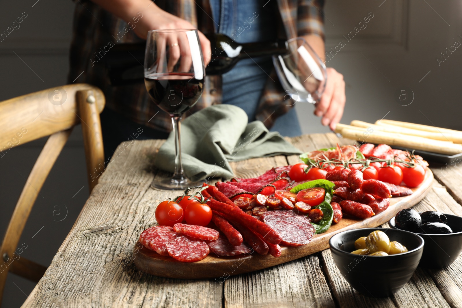 Photo of Woman pouring wine into glass at wooden table with different smoked sausages, selective focus