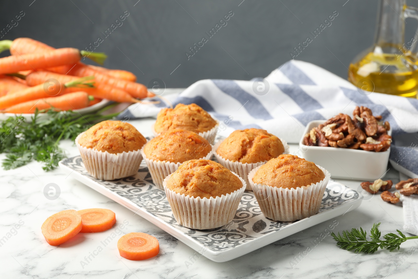 Photo of Tasty carrot muffins, fresh vegetables and walnuts on white marble table, closeup