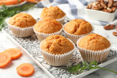 Photo of Tasty carrot muffins, cut vegetable and walnuts on white marble table, closeup