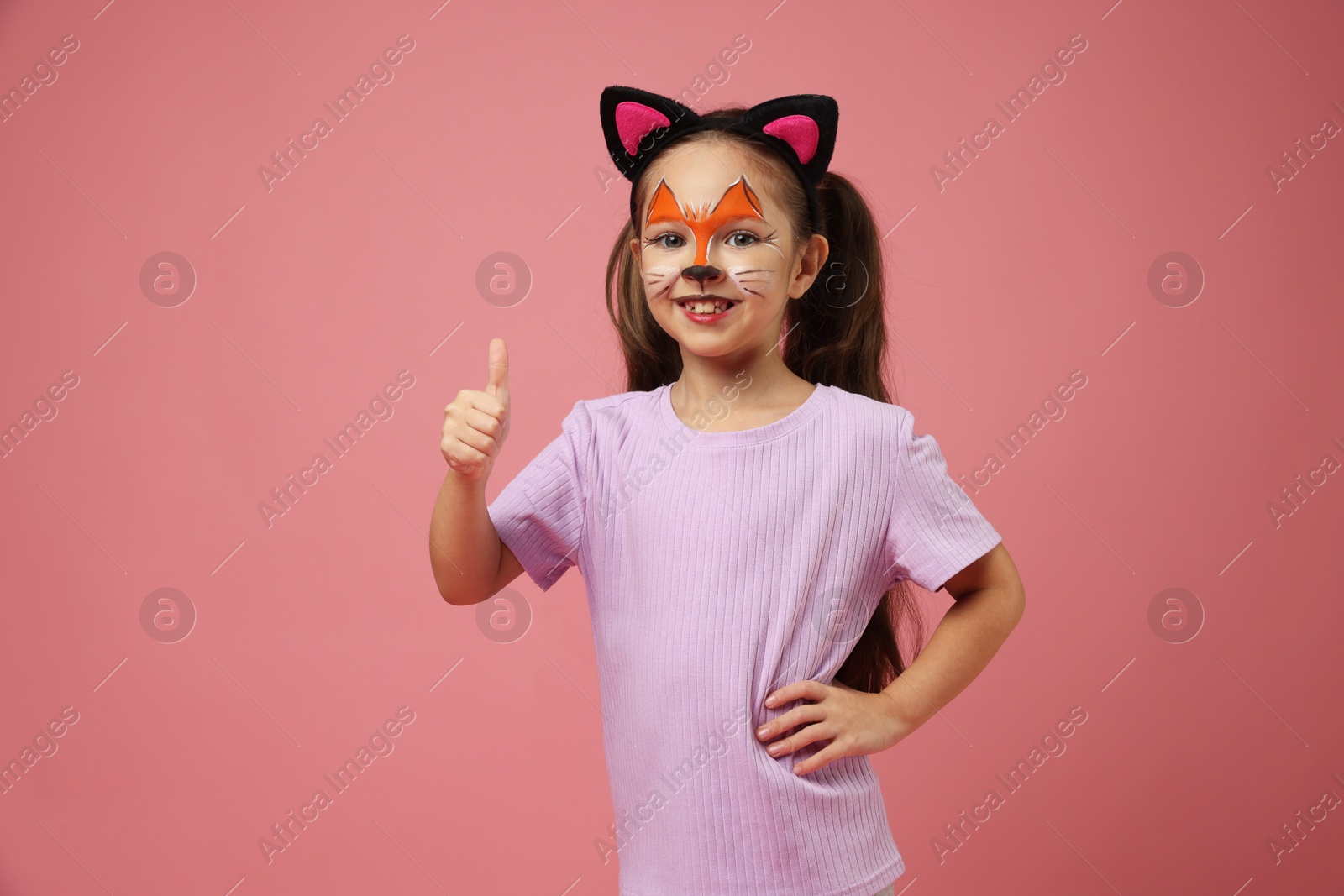 Photo of Cute girl with painted face and ears as cat showing thumbs up on pink background