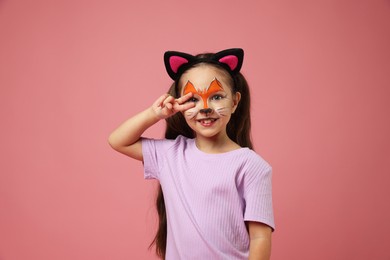 Photo of Cute girl with painted face and ears as cat showing peace sign on pink background