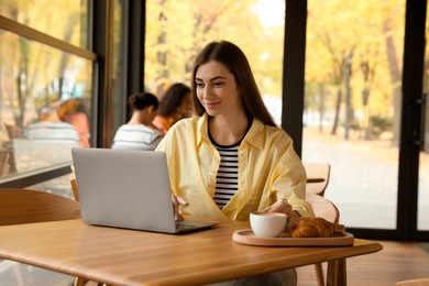 Photo of Woman with coffee and croissant working on laptop at table in cafe