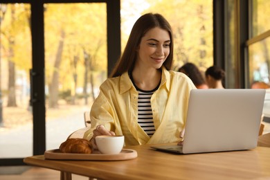 Photo of Woman with coffee and croissant working on laptop at table in cafe