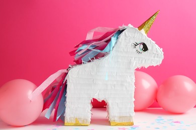 Photo of Bright pinata in shape of unicorn and balloons on white table against pink background