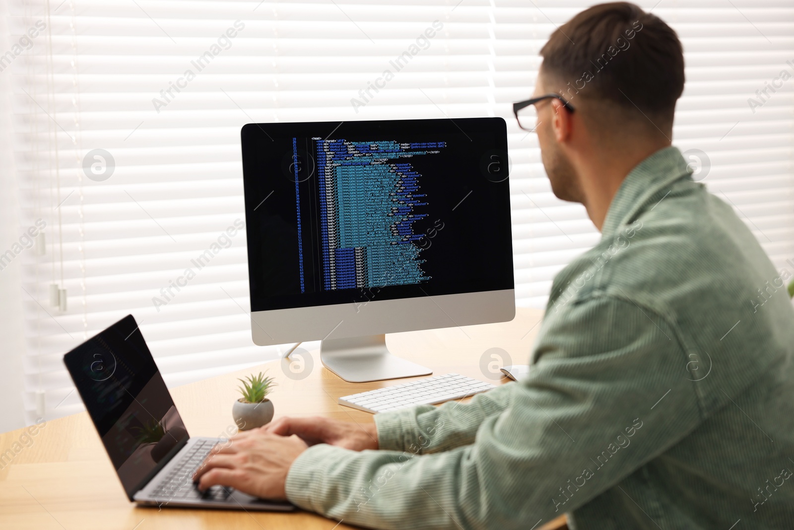 Photo of Programmer working on laptop and computer at wooden desk indoors