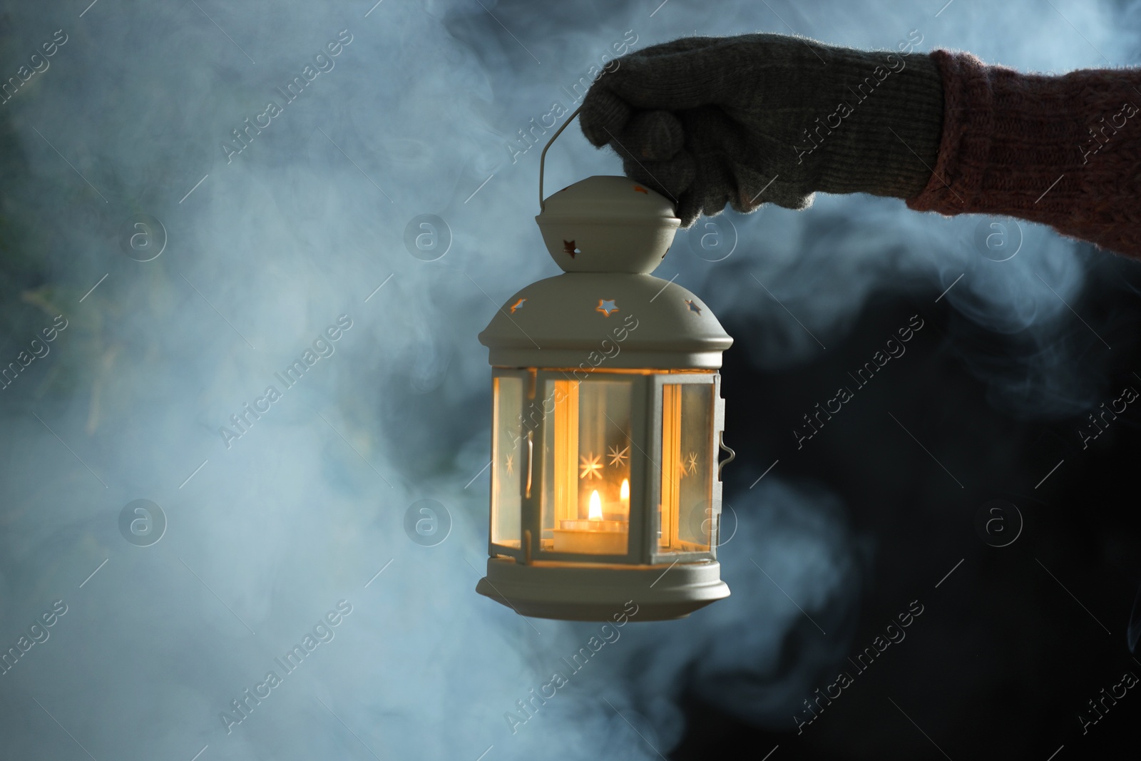 Photo of Woman holding Christmas lantern with burning candle on dark background with fog, closeup