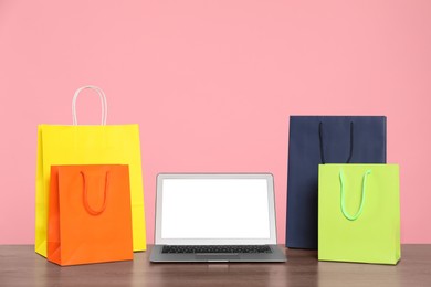 Photo of Internet shopping. Laptop and colorful paper bags on wooden table against pink background