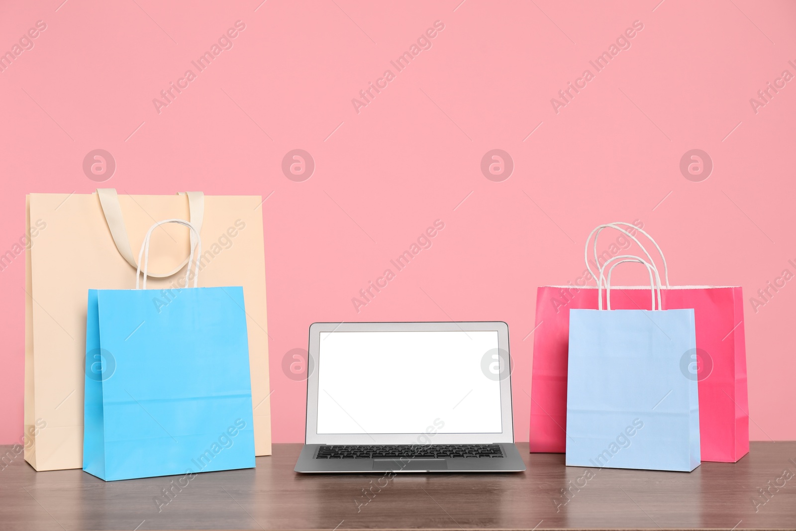 Photo of Internet shopping. Laptop and colorful paper bags on wooden table against pink background