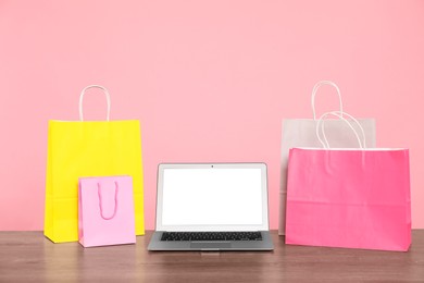 Photo of Internet shopping. Laptop and colorful paper bags on wooden table against pink background