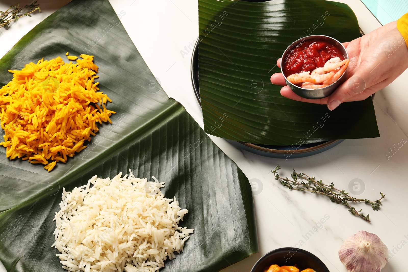 Photo of Woman holding bowl of shrimps and sauce at white table with banana leaves, above view