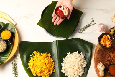 Photo of Woman holding bowl of shrimps and sauce at white table with banana leaves, top view