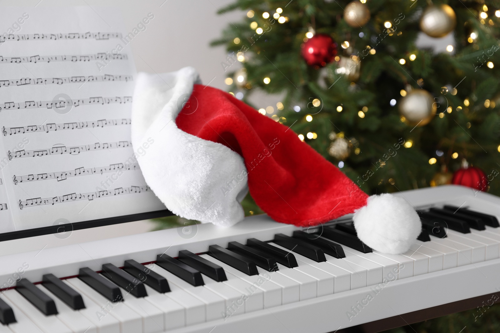 Photo of Synthesizer with music sheets and Santa hat near Christmas tree indoors, closeup