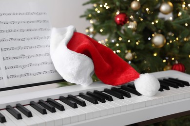 Photo of Synthesizer with music sheets and Santa hat near Christmas tree indoors, closeup