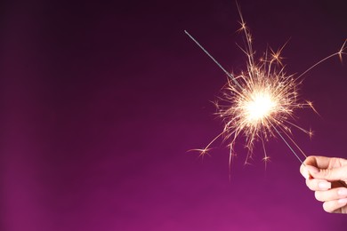 Photo of Woman with burning sparkler on purple background, closeup. Space for text