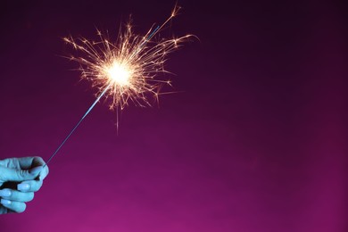 Photo of Woman with burning sparkler on purple background, closeup. Space for text