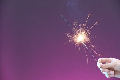 Photo of Woman with burning sparkler on violet background, closeup. Space for text