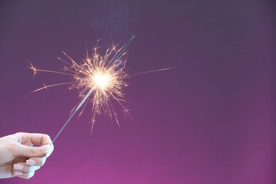 Photo of Woman with burning sparkler on violet background, closeup. Space for text