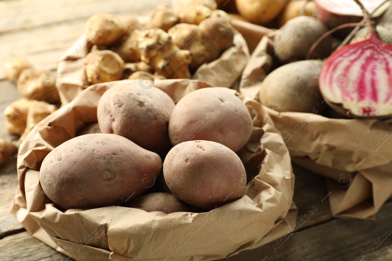 Photo of Different raw root vegetables in paper bags on wooden table, closeup