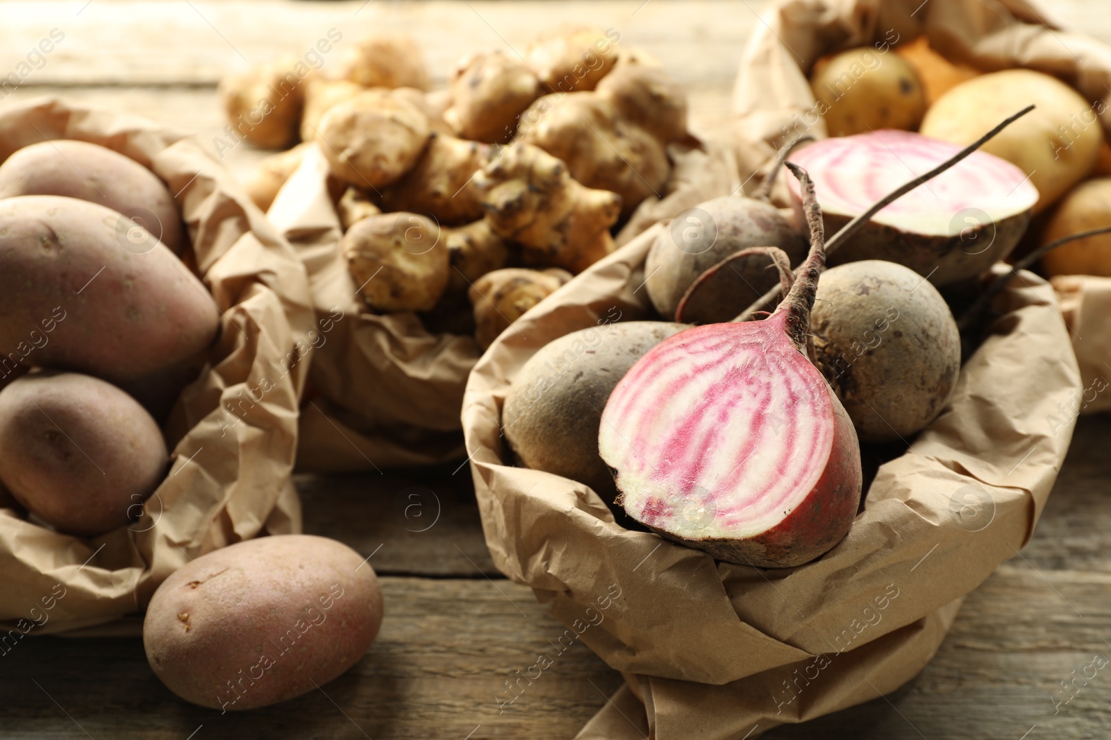 Photo of Different raw root vegetables in paper bags on wooden table, closeup