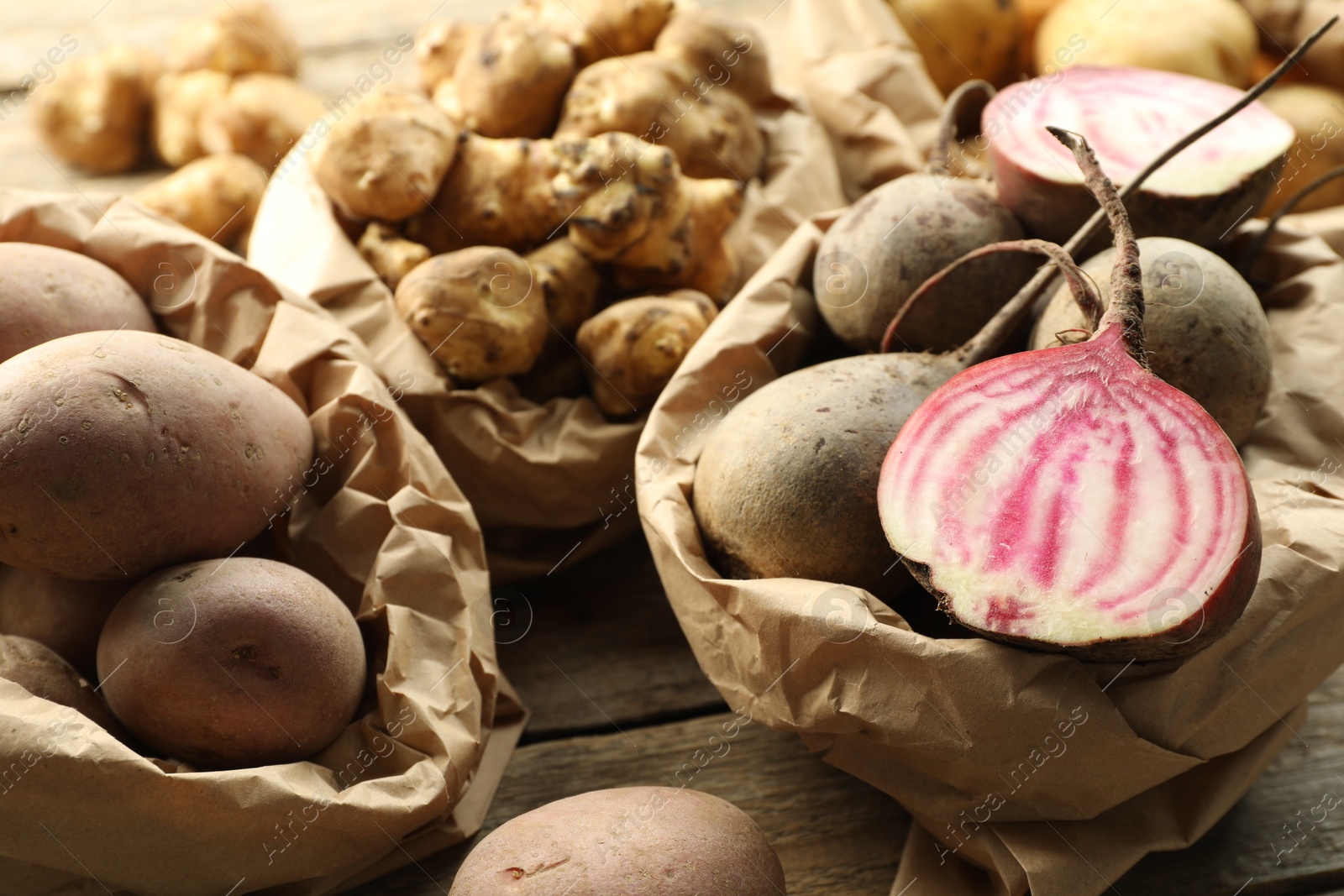 Photo of Different raw root vegetables in paper bags on wooden table, closeup