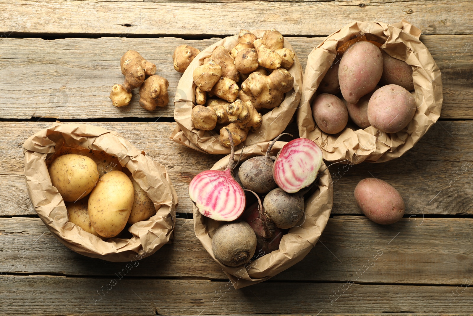 Photo of Different raw root vegetables in paper bags on wooden table, flat lay