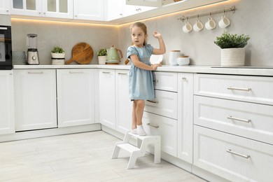 Little girl standing on step stool and reaching towards counter in kitchen