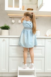 Little girl standing on step stool and reaching towards counter in kitchen, back view