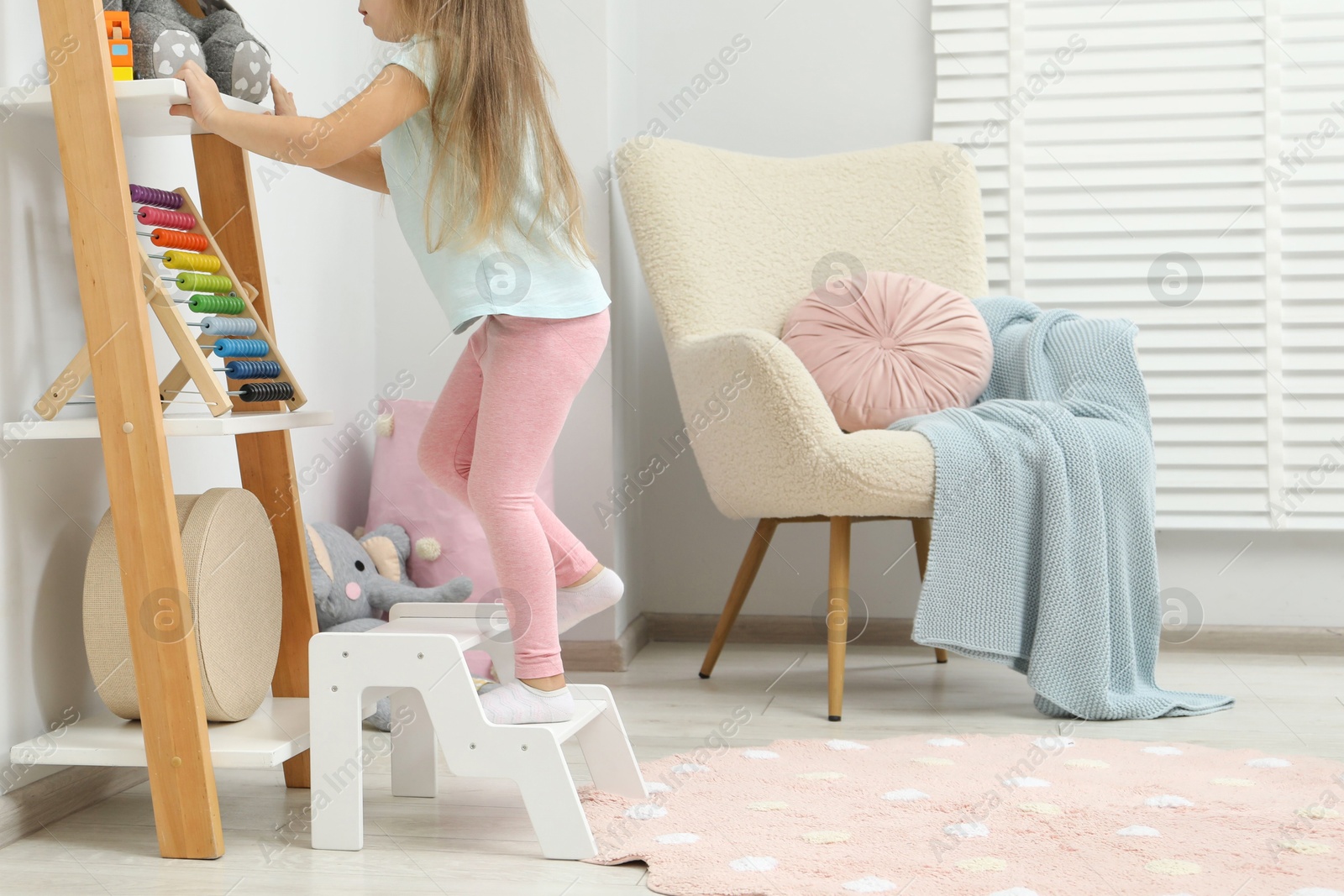 Photo of Little girl standing on step stool and reaching for toys on shelf indoors