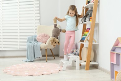 Photo of Little girl standing on step stool and reaching for toys on shelf indoors