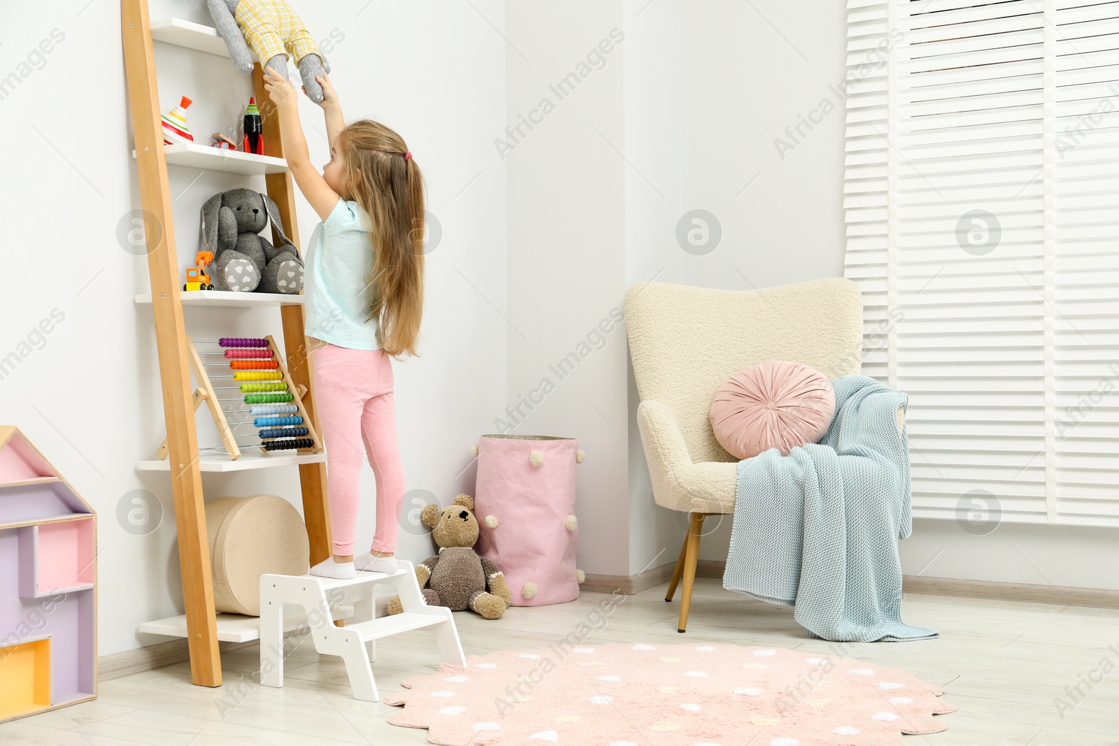 Photo of Little girl standing on step stool and reaching for toys on shelf indoors. Space for text