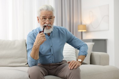 Photo of Handsome bearded man with tobacco pipe on sofa indoors