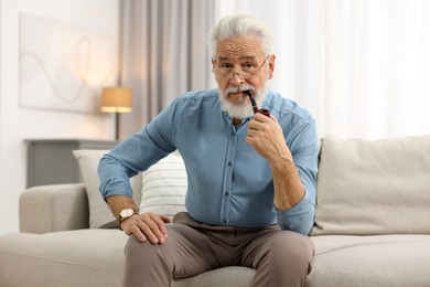 Photo of Handsome bearded man with tobacco pipe on sofa indoors