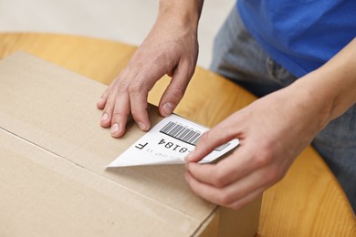 Photo of Man sticking shipping label with barcode on parcel at wooden table indoors, closeup