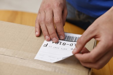 Man sticking shipping label with barcode on parcel at table indoors, closeup