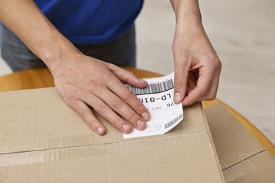 Photo of Man sticking shipping label with barcode on parcel at table indoors, closeup