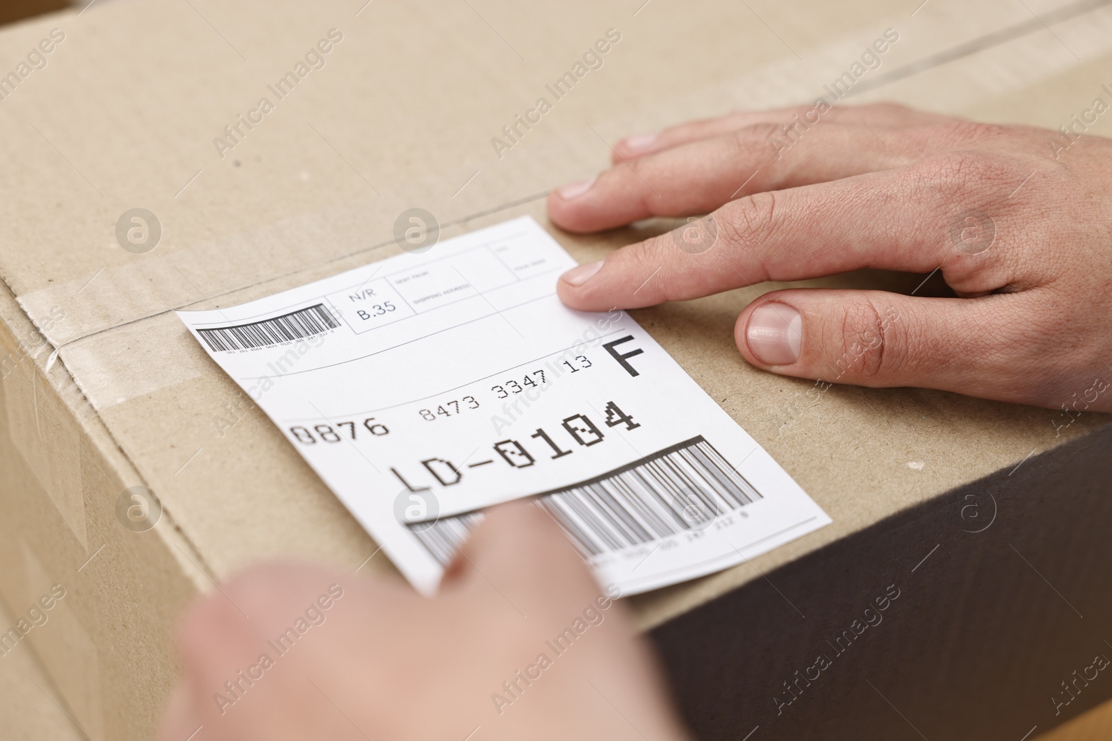 Photo of Man sticking shipping label with barcode on parcel indoors, closeup