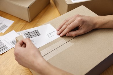 Photo of Man sticking shipping label with barcode on parcel at wooden table indoors, closeup