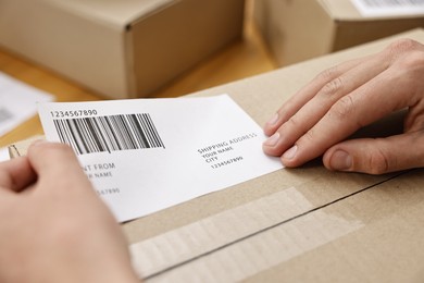 Photo of Man sticking shipping label with barcode on parcel indoors, closeup